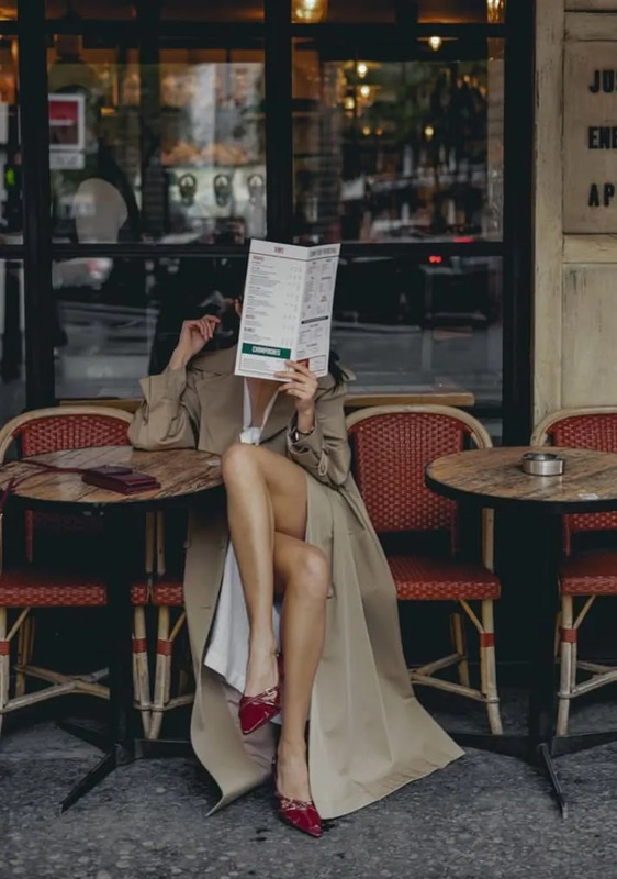 a woman sitting on a bench reading a paper in front of a restaurant with red chairs