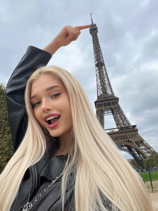a woman with long blonde hair standing in front of the eiffel tower holding her hand up