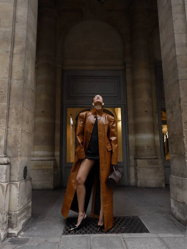 a woman standing in front of a building wearing a long brown coat and black dress