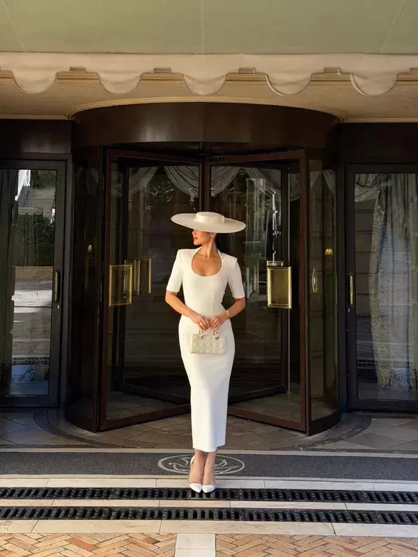 a woman in a white dress and hat is standing outside the entrance to a building