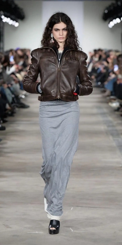 a woman walking down a runway wearing a brown jacket and grey skirt with white shoes
