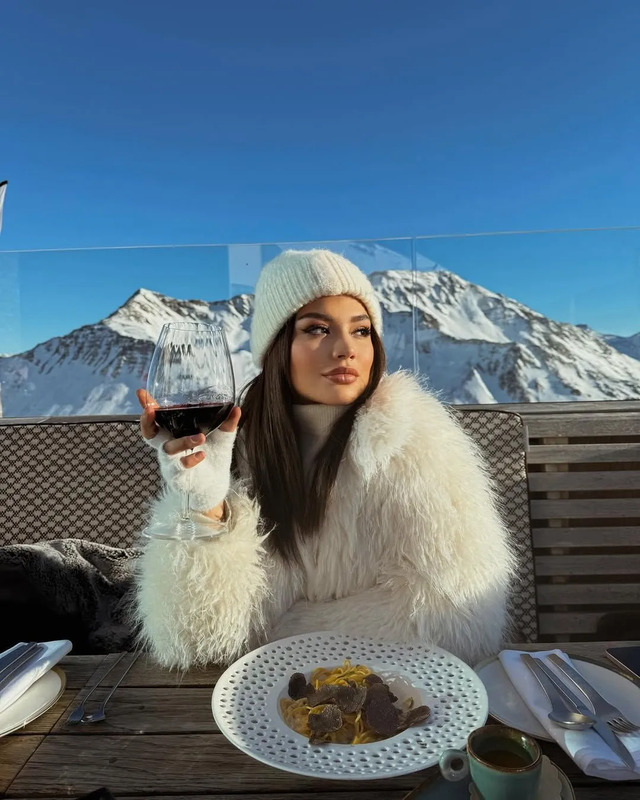 a woman sitting at a table with a glass of wine in front of her and mountains behind her