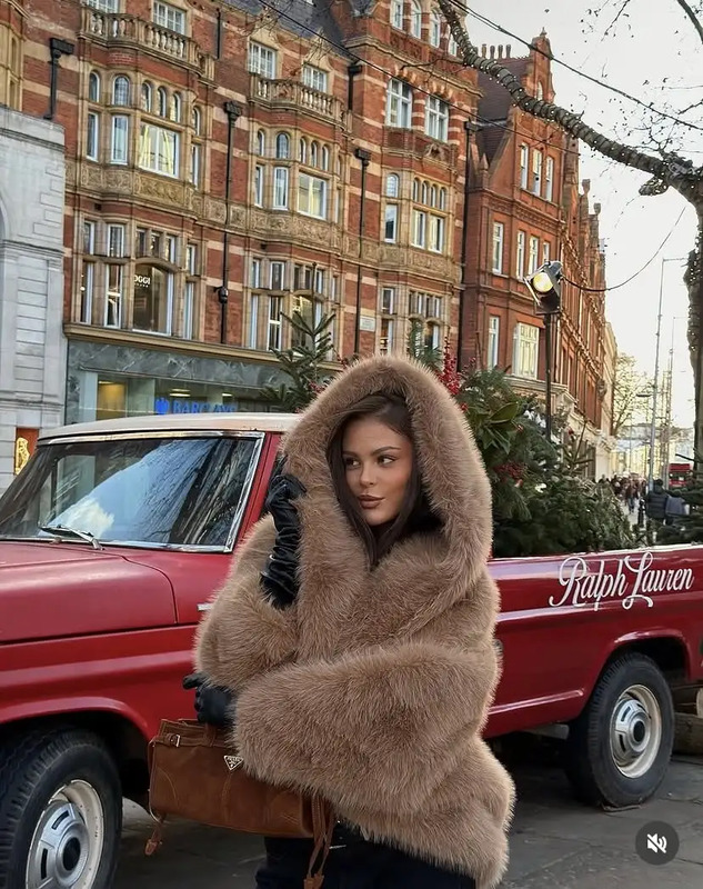 a woman in a fur coat is standing next to a red car and holding a suitcase