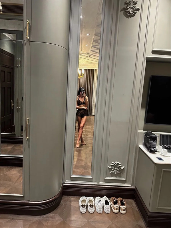 a woman taking a selfie in a bathroom mirror with shoes on the floor next to it