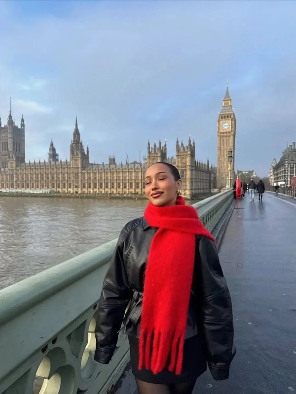 a woman wearing a red scarf and black jacket standing on a bridge near the water