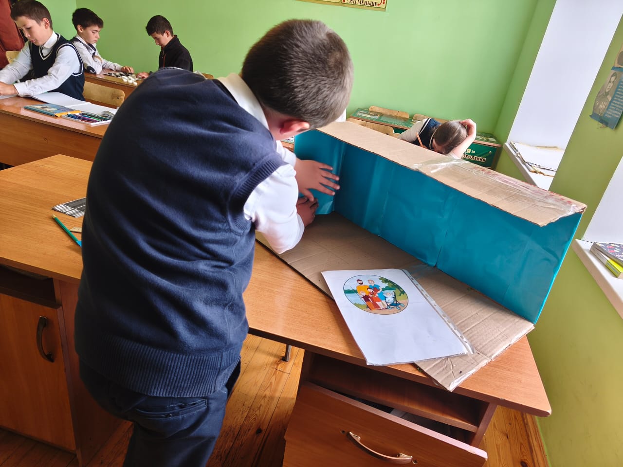 Небо — символ большого пути, свободы и высоты, к которой стремится Ахмади. Оно визуально связывает прошлое и будущее: по «взлётной полосе» зритель идёт от фото самолётов старших поколений к самолёту мечты мальчика, а над всем этим — общее небо, которое словно говорит: традиция продолжается.
Заклеивая картон, мальчик учился работать с материалами, подбирать цвет, аккуратно наносить клей и разглаживать бумагу — то есть осваивал навыки ручного труда. Педагог помогал выбрать оттенок и следить за тем, чтобы фон был ровным и без пузырей.
Так простая техническая деталь становится выразительным элементом, который усиливает главную мысль работы: уважение к прошлому и стремление к будущему.