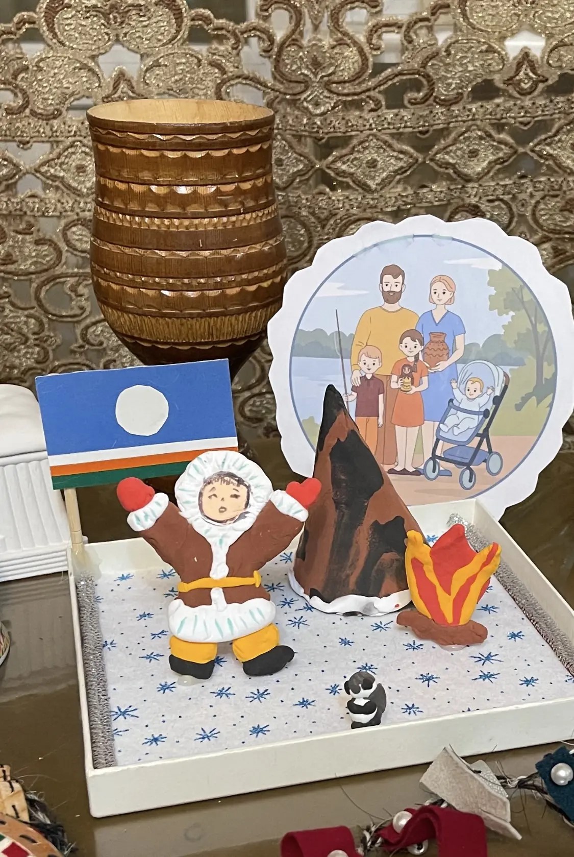 Моя творческая композиция для конкурса «Моя семья — моя Россия», выполненная в смешанной технике с активным использованием пластилина В центре — фигурка ребёнка в традиционной северной одежде: объёмная лепка передаёт фактуру меховой парки, варежек и унтов. Рядом — чум и яркий костёр: рельеф пламени и тёмные слои чума созданы из пластилина разных оттенков. У подножия чума — маленький пингвиненок, тоже вылепленный из пластилина. Слева — наш миниатюрный флаг Якутии с кругом и цветными полосами, это аппликация, дополняющая сюжет о семейных традициях. На заднем плане — деревянный чорон, традиционный якутский сосуд для кумыса: его резной орнамент и слоистая фактура подчёркивают национальный колорит работы. Вся сцена размещена на подложке с узором из снежинок — это задаёт зимнюю северную атмосферу. Моя работа объединяет символы малой родины: природу, быт, семейные ценности и культуру народа саха.