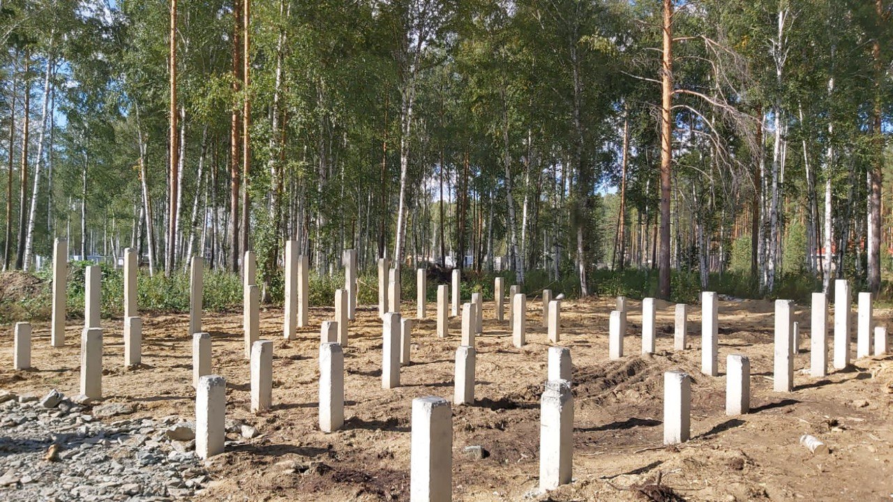 Свайное поле из железобетонных стоек на подготовленной площадке в лесу, справа видна жёлтая строительная техника