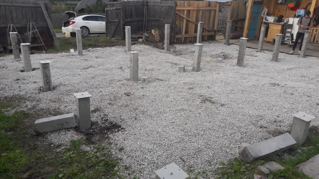 Свайно-столбчатый фундамент на слое щебня перед деревянной верандой; установленные бетонные столбы, в кадре человек.