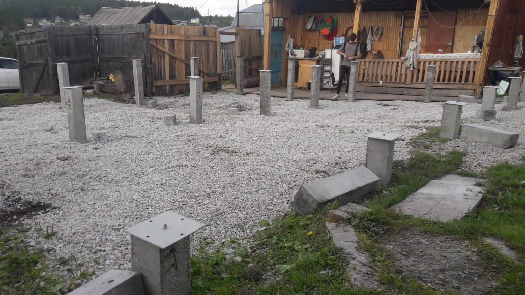 Свайно-столбчатый фундамент: бетонные столбы на щебёночной подушке во дворе; на заднем плане деревянный забор и белый автомобиль.