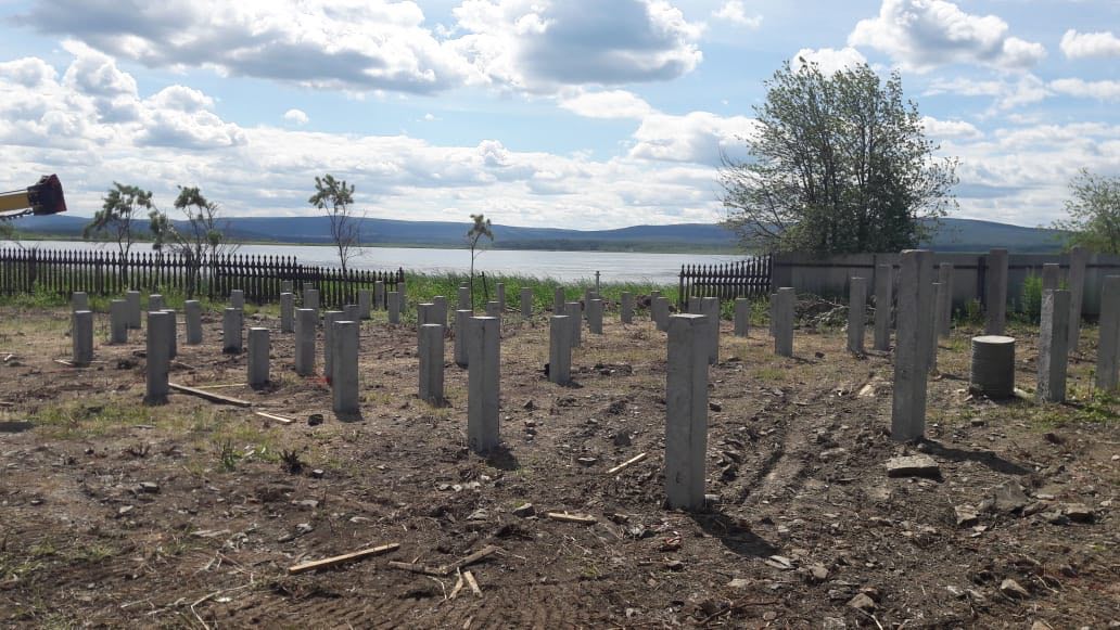 Свайное поле на подготовленной площадке; на заднем плане бытовка, забор и деревья.