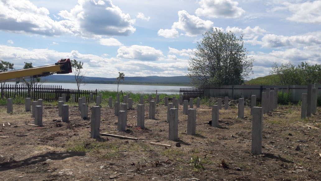 Железобетонные сваи под фундамент коттеджа, вид с правой стороны; забор, деревья и частные дома у озера.