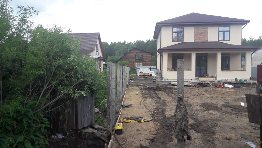 Фундамент под забор с бетонными стойками; кустарник справа, в конце линии опор виден рабочий, на земле строительные материалы (Нижний Тагил, Черемушки)