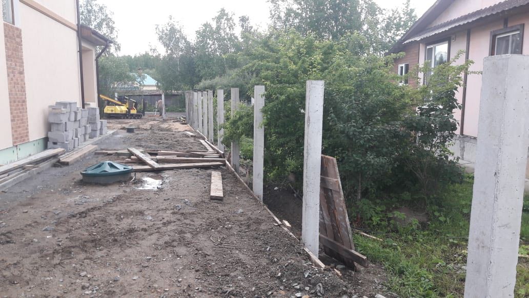 Монтаж бетонных столбов под забор у частного дома: вид вдоль фундамента, блоки и доски слева, густые кусты справа (Нижний Тагил, Черемушки)