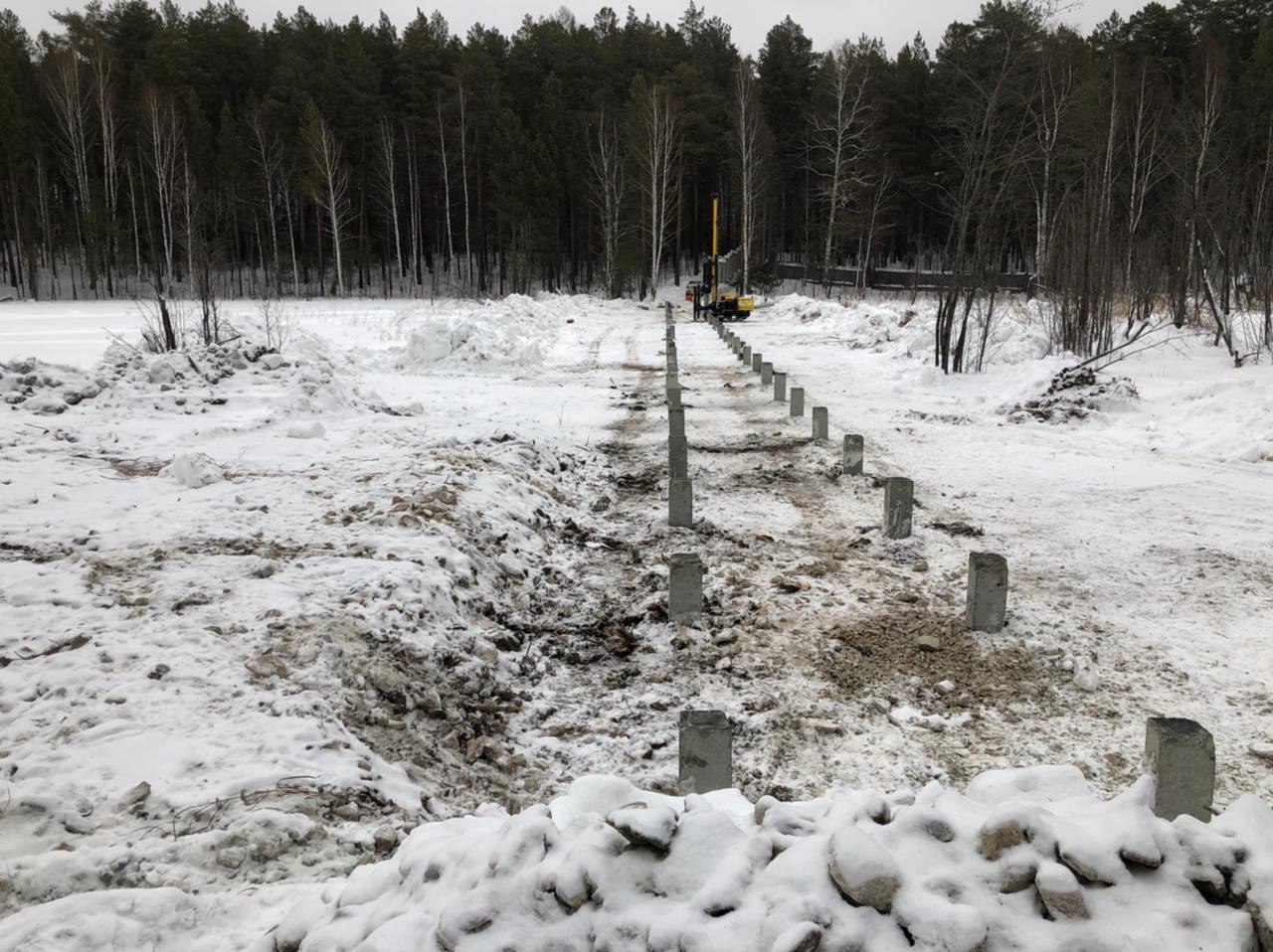 Ряды оголовков свай вдоль стены здания; на снегу следы техники и лежат балки.