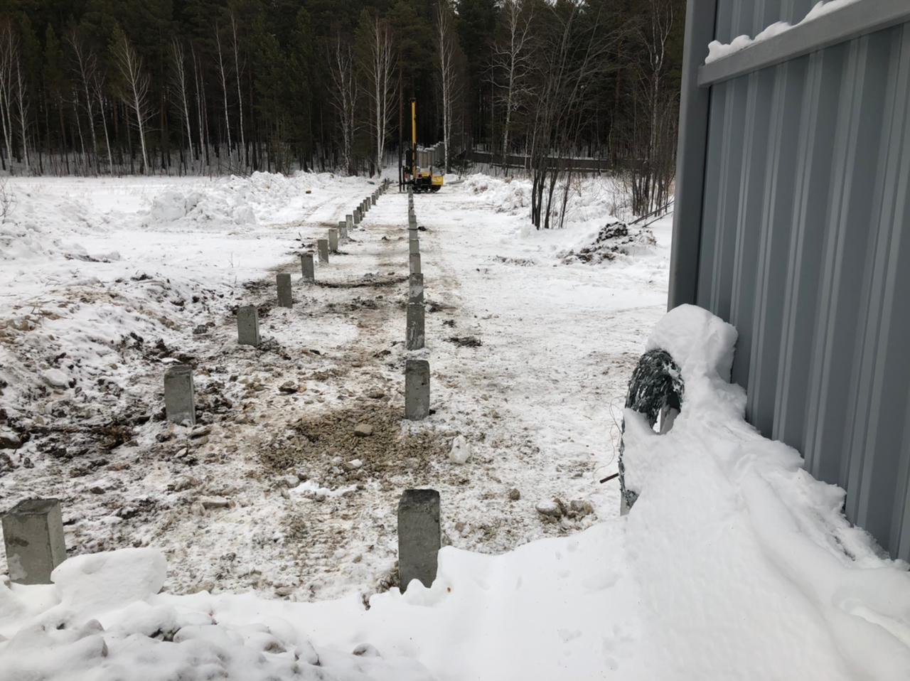 Две параллельные линии ЖБ свай на заснеженном участке, вдали кран/сваебойная установка на фоне леса.