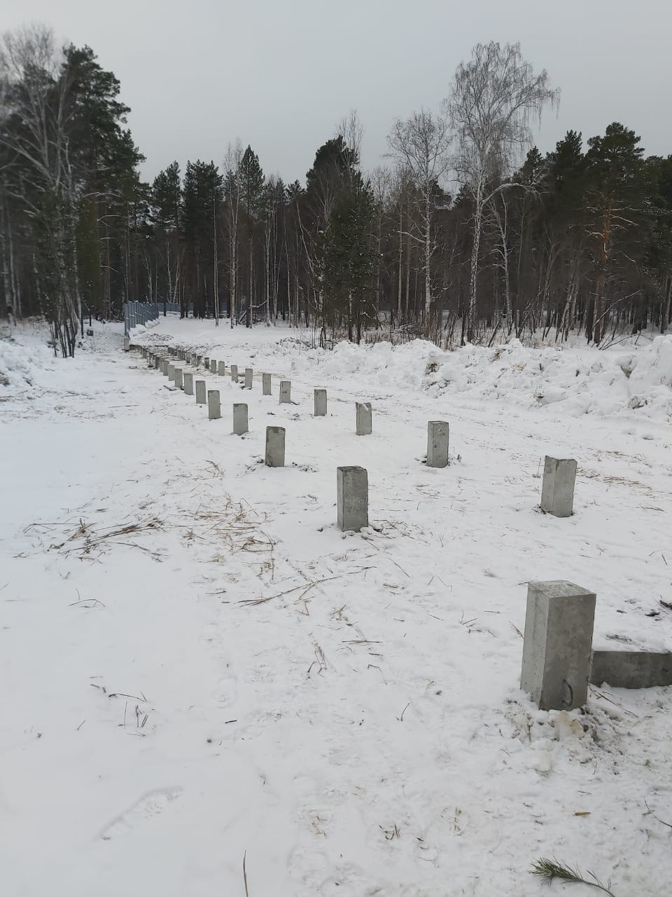 Несколько оголовков свай у торца металлического здания; сугробы и лесная кромка на фоне.