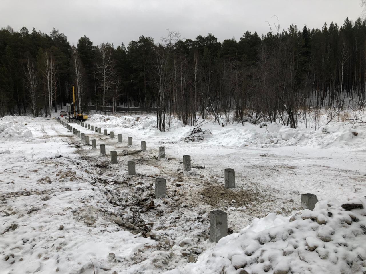 Ряды железобетонных свай вдоль стены здания на зимней стройплощадке; на горизонте лес и техника.