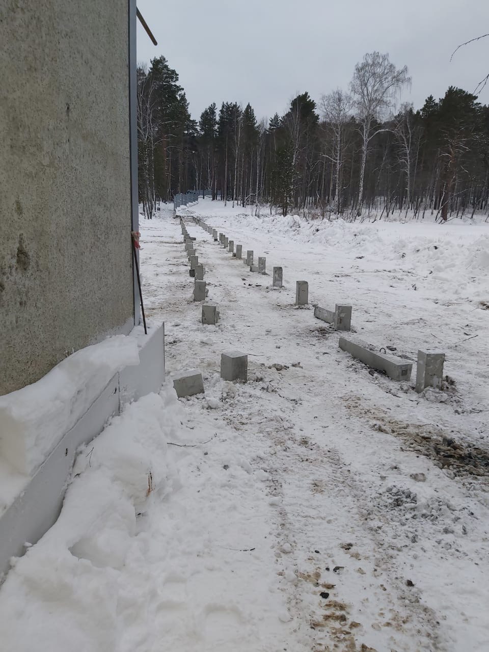 Ряд забитых железобетонных свай на заснеженной площадке; вдали видна сваебойная установка и лес.